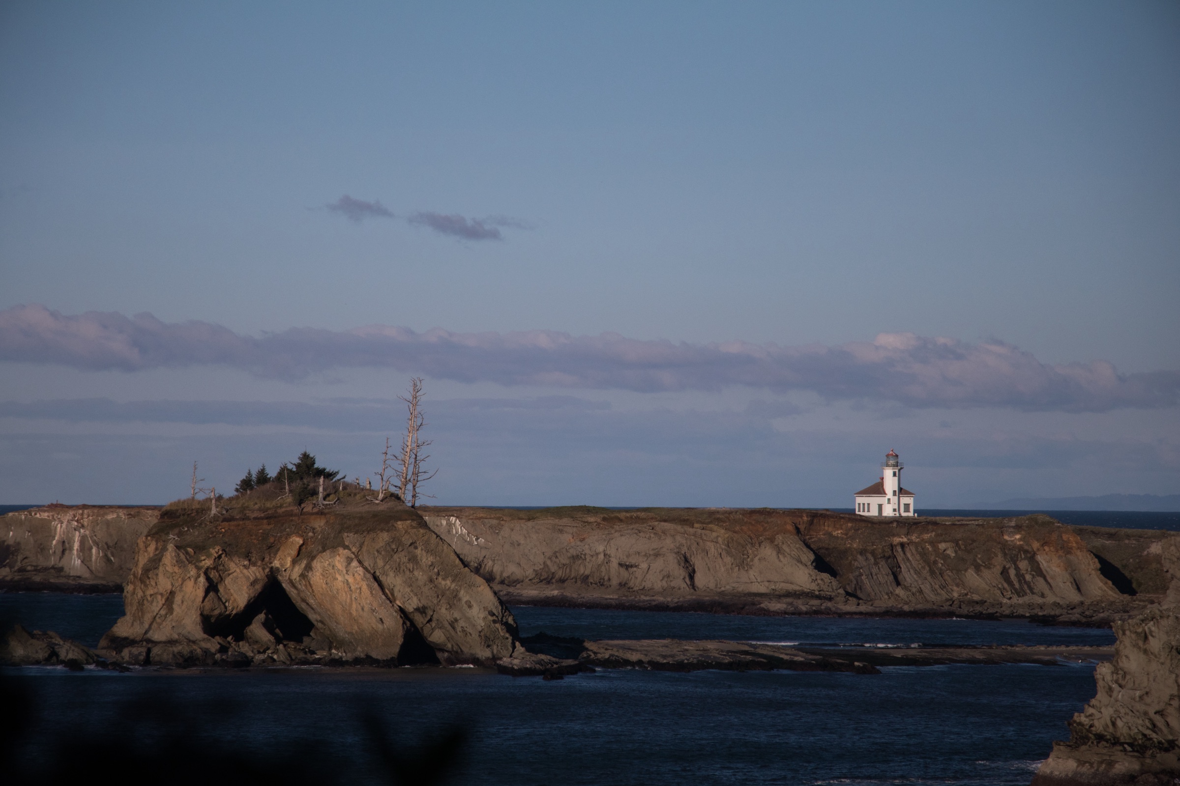 A lighthouse on a rocky coastline beneath a blue evening sky.